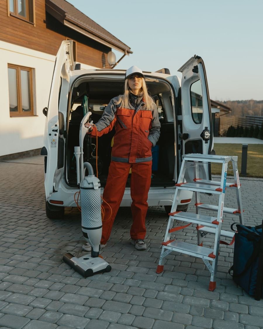 Cleaning professional in orange overalls standing at the open back of a van, with a vacuum cleaner and stepladder ready