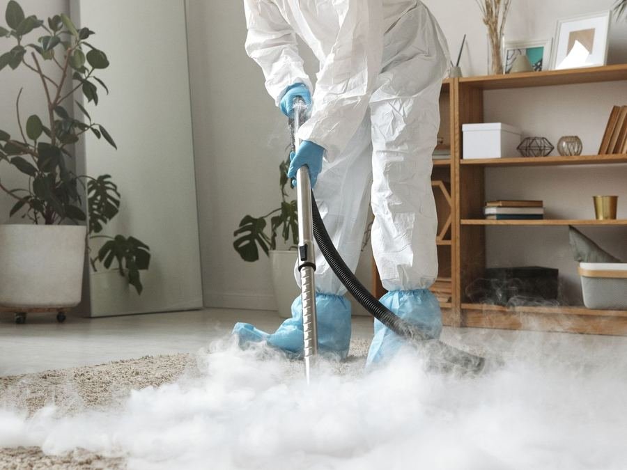 Cleaner in a protective suit and blue gloves using a vacuum wand on a carpet in a cloud of steam
