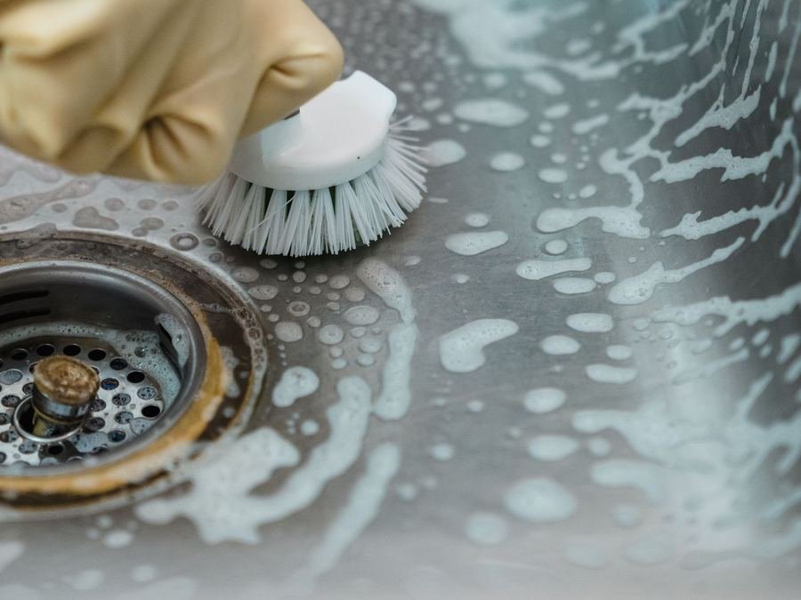 Gloved hand scrubbing a soapy stainless-steel sink and drain with a small brush