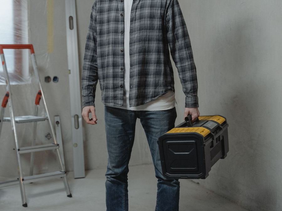 Handyman carrying a toolbox through a room under renovation, with a stepladder behind him