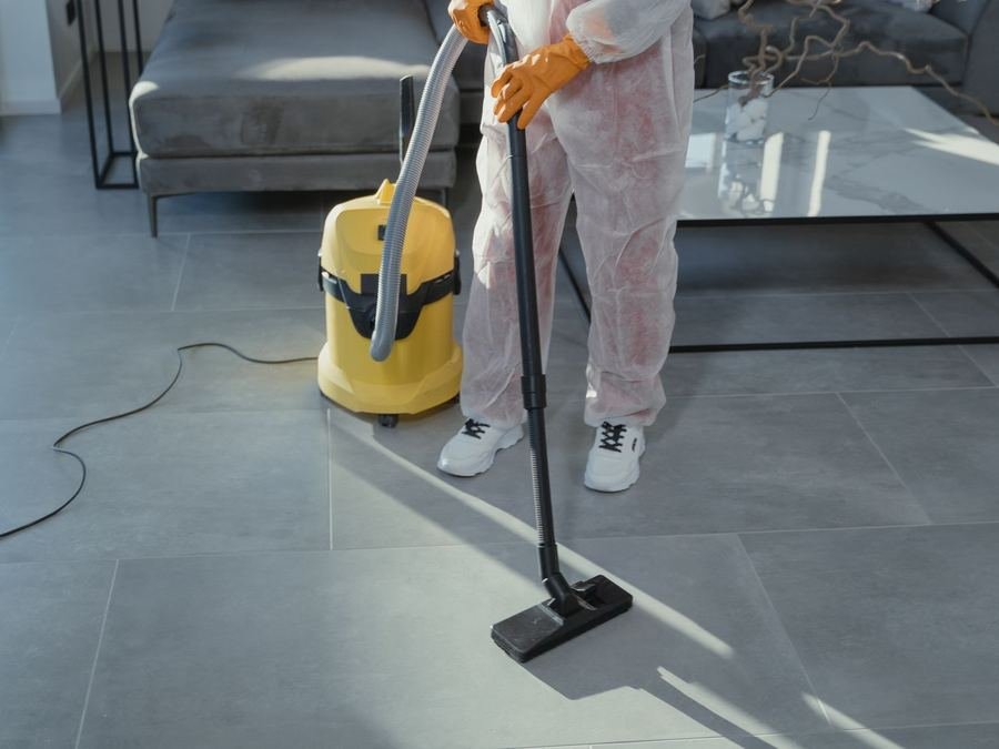 Cleaner in protective overalls and orange gloves vacuuming a tiled living-room floor
