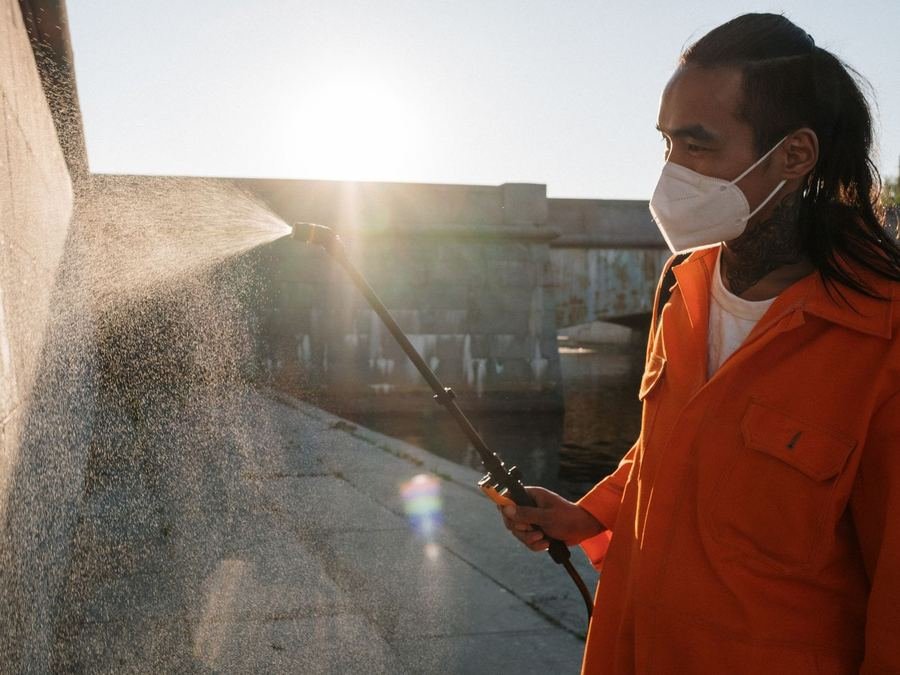 Worker in an orange overall and face mask spraying a wall and pavement outdoors with a spray wand