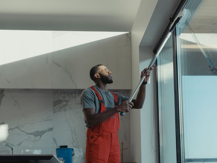 Window cleaner in orange overalls using an extension pole to wash a tall window in a modern home
